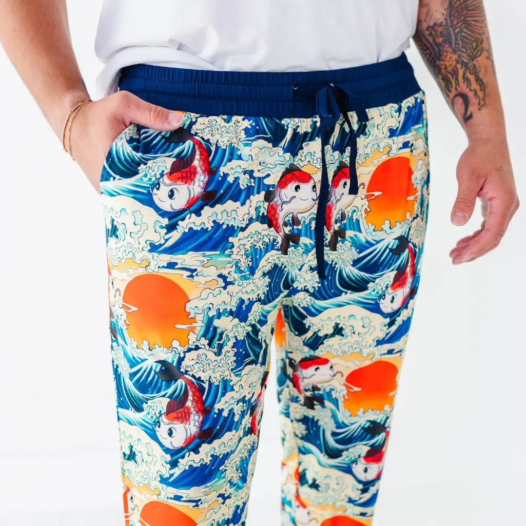 Close-up of a man wearing pajama pants with a koi fish, ocean wave, and sunset print and a navy drawstring waistband.